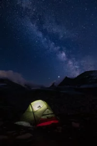 Tente éclairée sous un ciel étoilé avec la Voie lactée visible au-dessus d’un paysage de haute montagne.