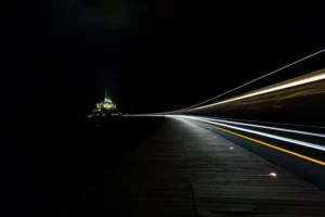 Pose longue de nuit avec lumières artificielles, traînées lumineuses des phares sur une route menant vers Le Mont-Saint-Michel.