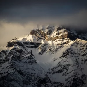 Sommet rocheux enneigé sous des nuages sombres, lumière dorée d’hiver